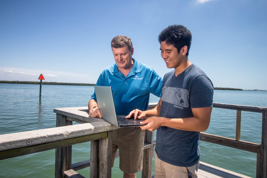 Professor and young man work on a laptop on a dock by the water.