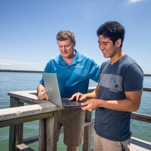 Professor and young man work on a laptop on a dock by the water.