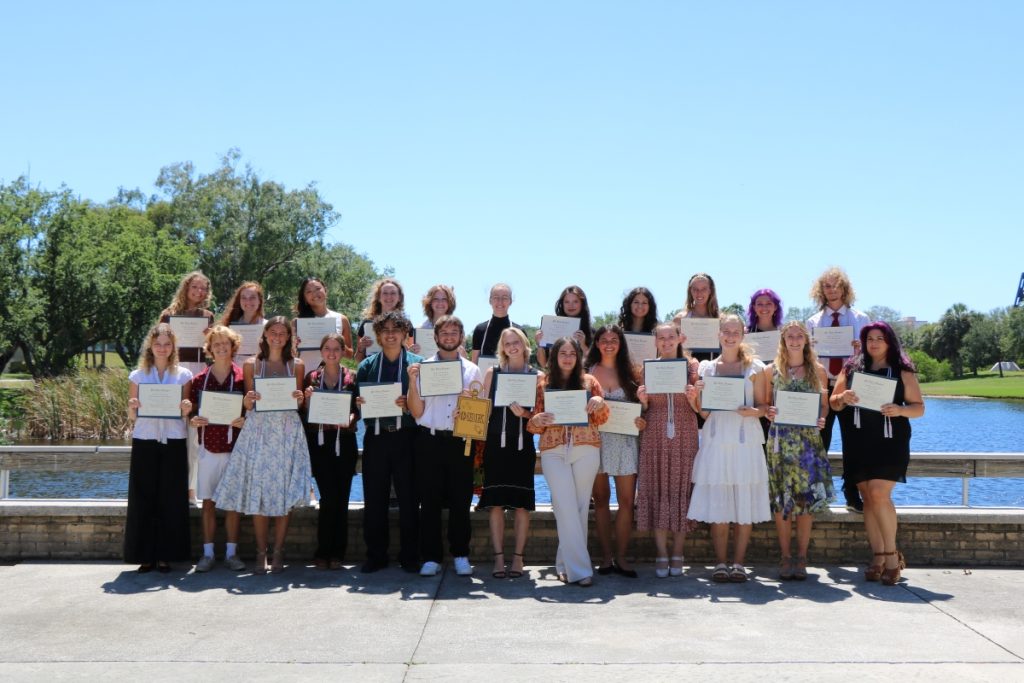 A group of students stand outside in front of a pond displaying framed certificates.
