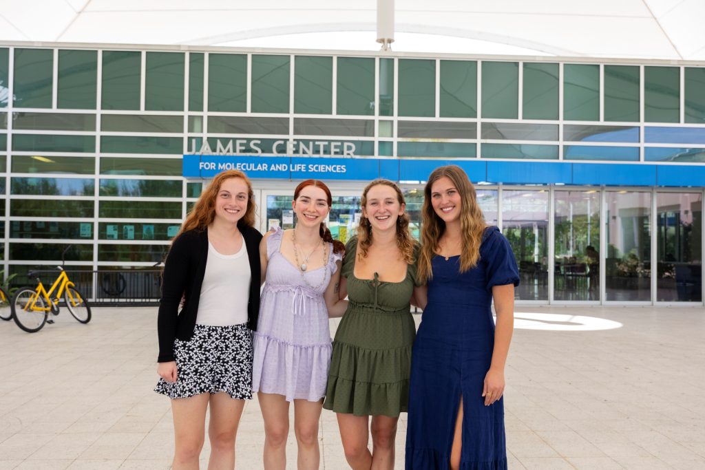 Four female Eckerd College students pose for a picture in front of a glass building.