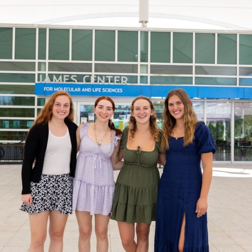 Four female Eckerd College students pose for a picture in front of a glass building.