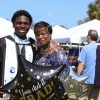 Student in regalia stands outside in front of a large tent with his mother while holding a balloon that reads YOU DID IT