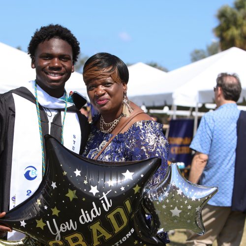 Student in regalia stands outside in front of a large tent with his mother while holding a balloon that reads YOU DID IT