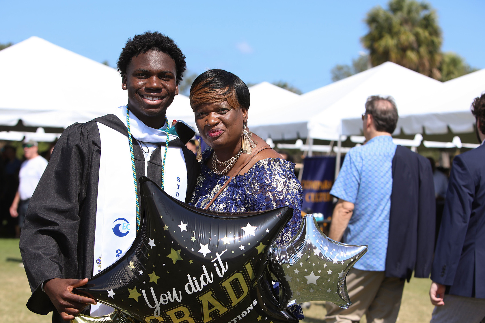 Student in regalia stands outside in front of a large tent with his mother while holding a balloon that reads YOU DID IT