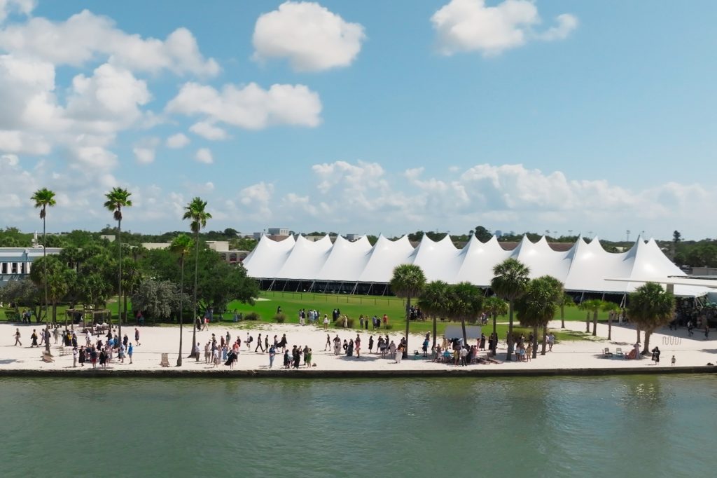 Large white tent set atop a grassy field and a seawall and a beach with dozens of people walking along