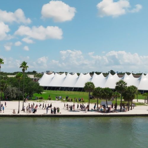 Large white tent set atop a grassy field and a seawall and a beach with dozens of people walking along