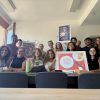 A group of around a dozen people sit at a table smiling and holding up a framed display.