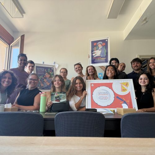 A group of around a dozen people sit at a table smiling and holding up a framed display.