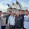Seven young men in baggy jackets and sweatshirts stand in front of the Sydney Opera House.