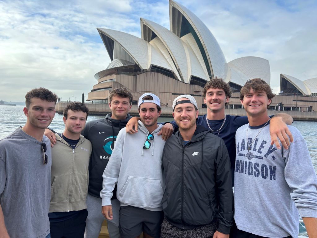 Seven young men in baggy jackets and sweatshirts stand in front of the Sydney Opera House.