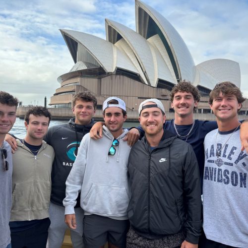 Seven young men in baggy jackets and sweatshirts stand in front of the Sydney Opera House.