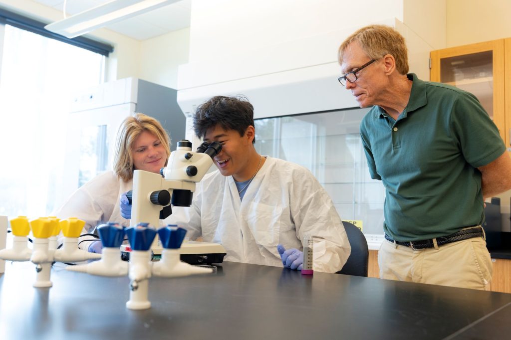 Allen Chen, in a white lab coat, looks into a microscope as a classmate and a professor look on.