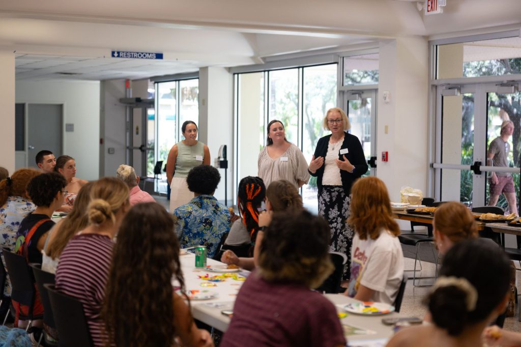 Three women stand at the front of a crowded room of seated students.