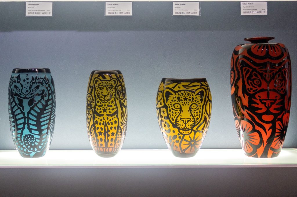 Four glass vases on a display shelf with tags identifying what each one is named on who made it affixed to the wall behind them.