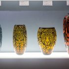 Four glass vases on a display shelf with tags identifying what each one is named on who made it affixed to the wall behind them.