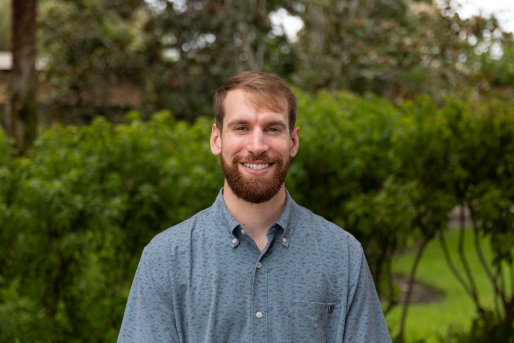 A smiling man standing in front of a background of green healthy shrubbery.