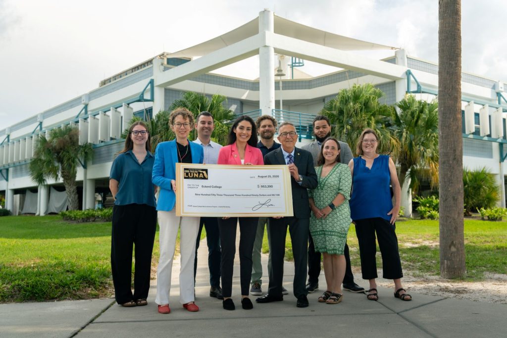 A group of nine people outdoors hold a giant check, and there is a large building in the background.