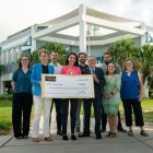 A group of nine people outdoors hold a giant check, and there is a large building in the background.