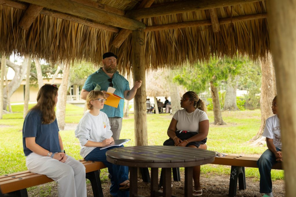 I man in a teal shirt speaks to four seated students in a thatched hut.