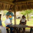 I man in a teal shirt speaks to four seated students in a thatched hut.