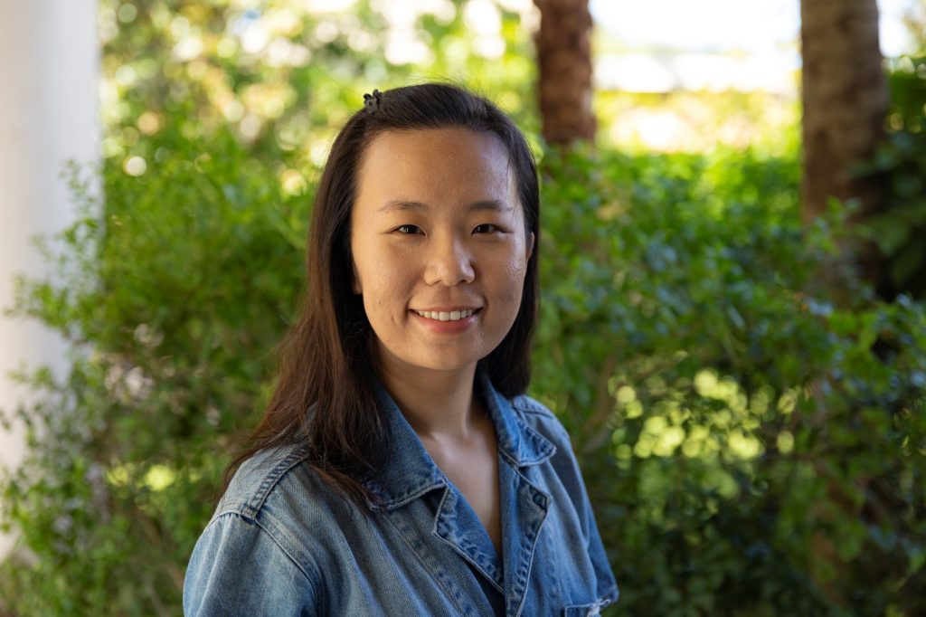 A woman in a blue denim shirt standing in front of a bush.