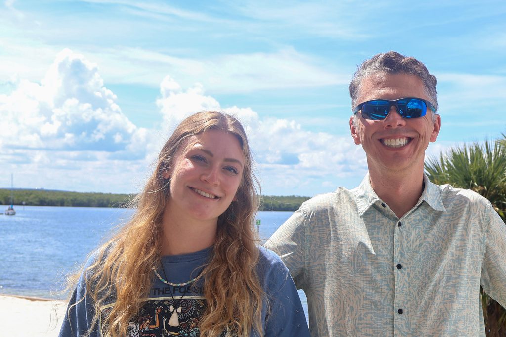 A student in a t-shirt and a gentleman in a collared shirt and sunglasses stand side by side smiling on a balcony overlooking a sandy beach and bay