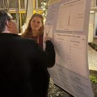 A person in a black suit jacket examines a poster as a young woman looks on.