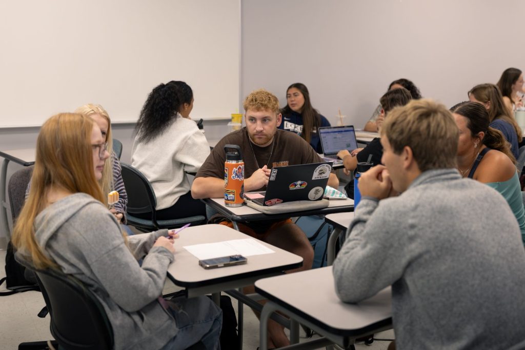 A group of men and women sit with their desks in circled arrangements of four each talking.
