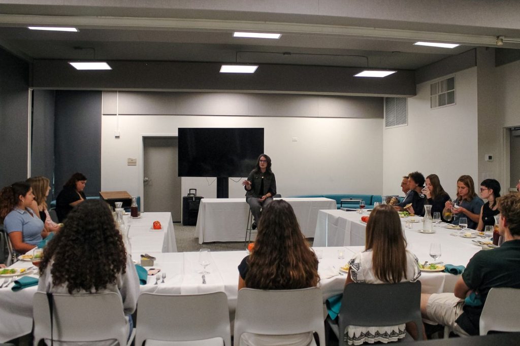 A woman sits at the front of a conference room speaking to a group of people seated at tables in a U shape around her.