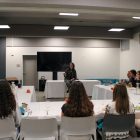 A woman sits at the front of a conference room speaking to a group of people seated at tables in a U shape around her.