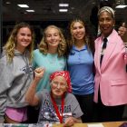 Four women stand behind one elderly woman in a red bandana who is seated and raising her fist.