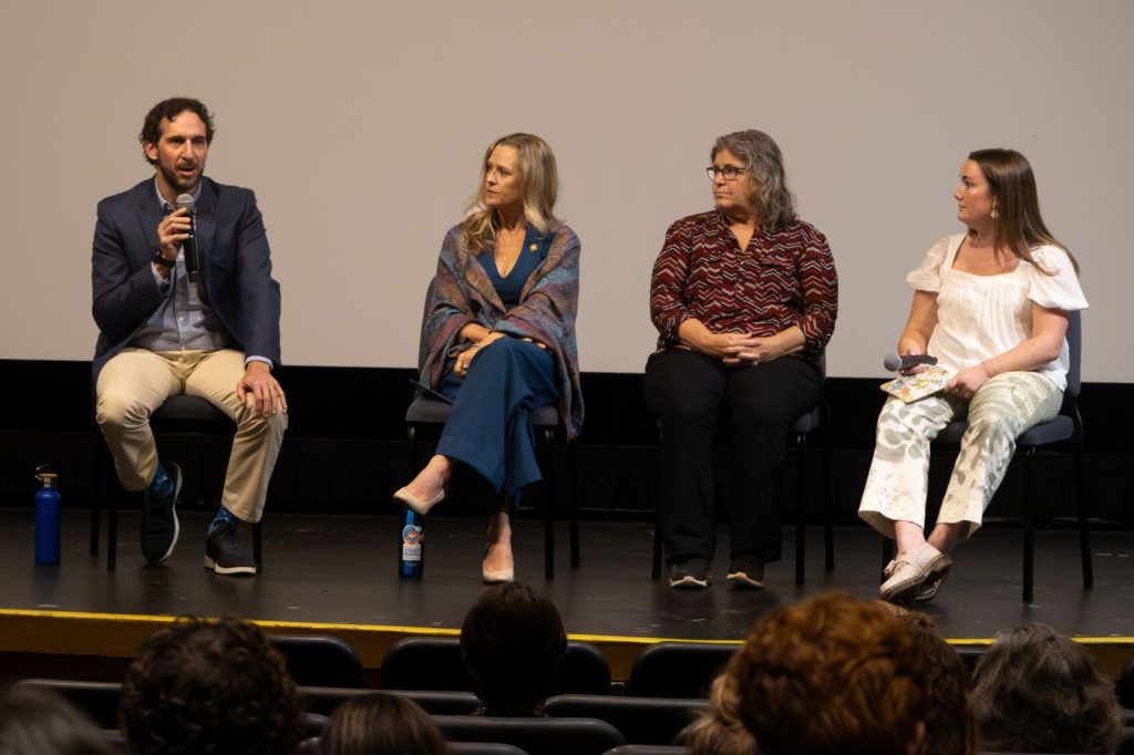 Four people sit on a stage and the one on the far left is speaking into a microphone.