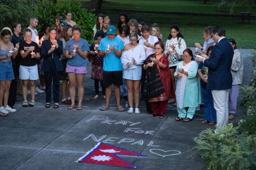 A group of people hold a candlelight vigil over a chalk drawing that reads "Pray for Nepal."