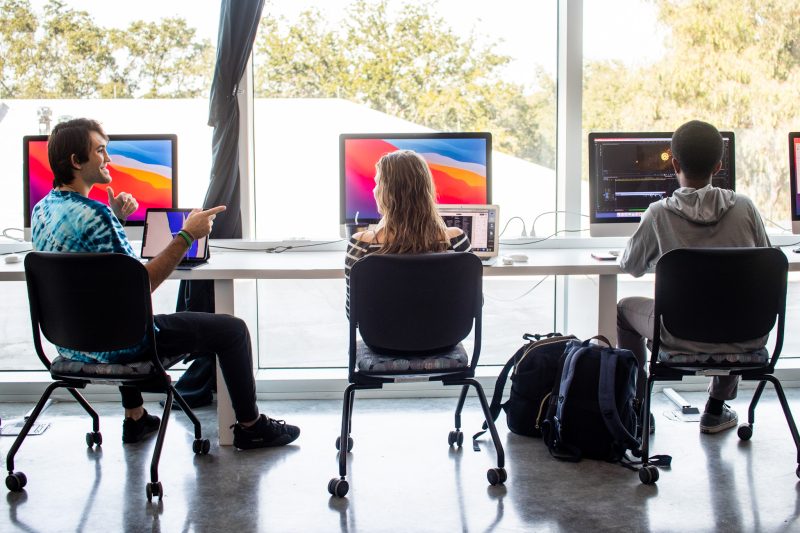 Three students seated side by side in front of colorful Mac computers and monitors by a big window overlooking a natural landscape