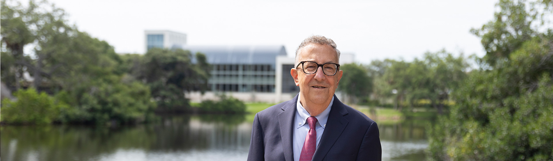 Man in suit stands before a pond with an academic building in the background