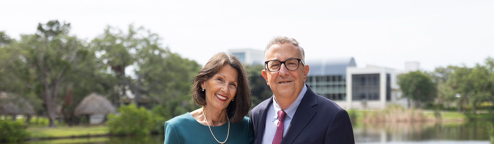 A couple in formal attire stands before a pond with an academic building in the background