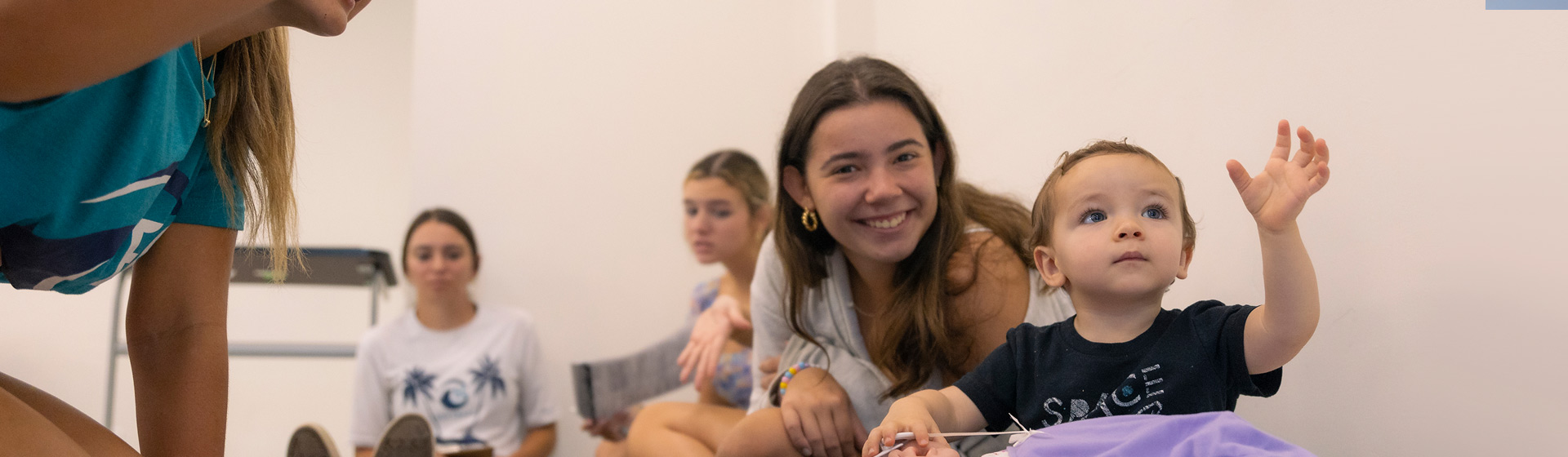 College students seated on the floor with a toddler who is sitting and pointing