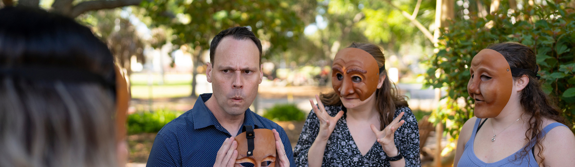 Professor imitates the face on a mask he holds and two students wearing masks stand next to him