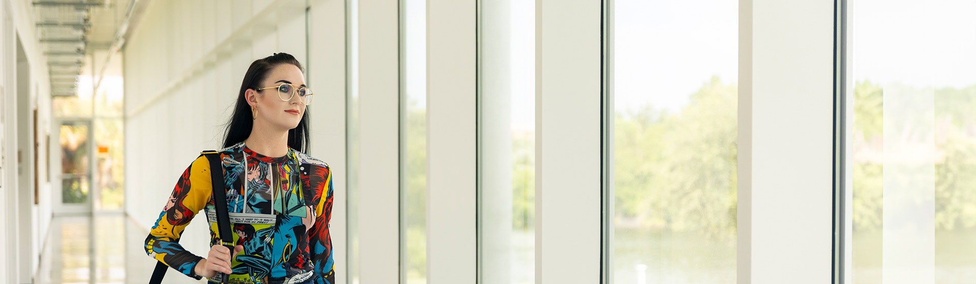 Woman in glasses and print shirt walking down bright hallway of windows with bag over shoulder