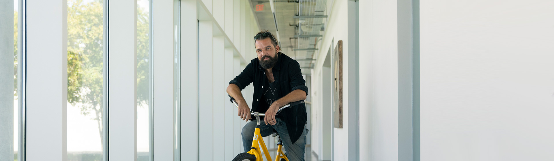 Professor with beard on yellow bike in a hallway lined with windows