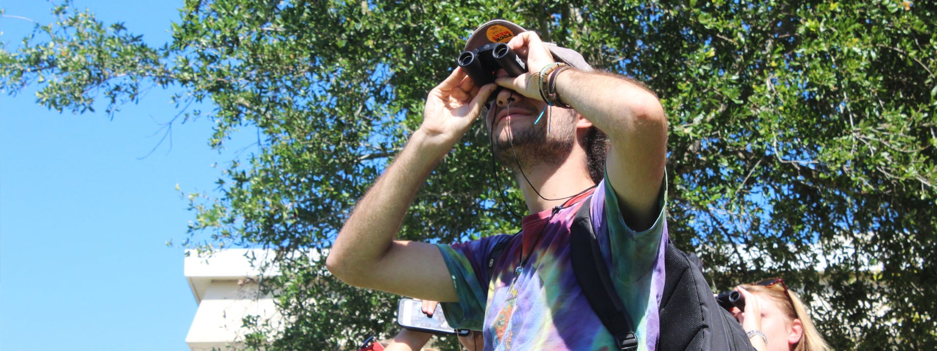 Male student in tie-dye shirt looking through binoculars
