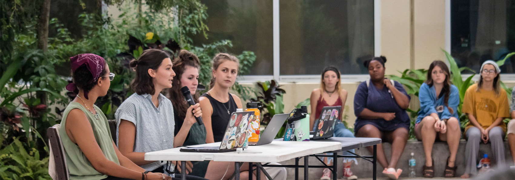 Four female students sit at desk and speak into microphone as audience listens