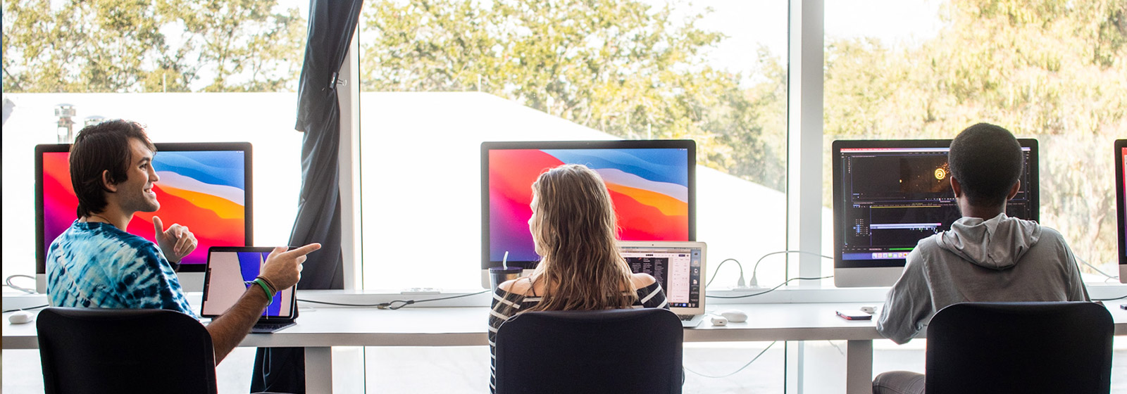 Students sitting in front of big Mac computers in a lab by window