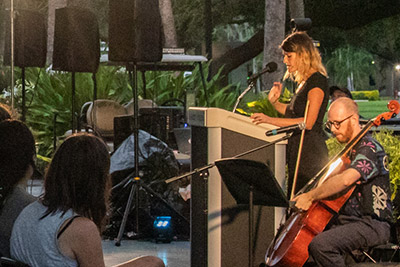 Woman speaks at podium while musician plays cello
