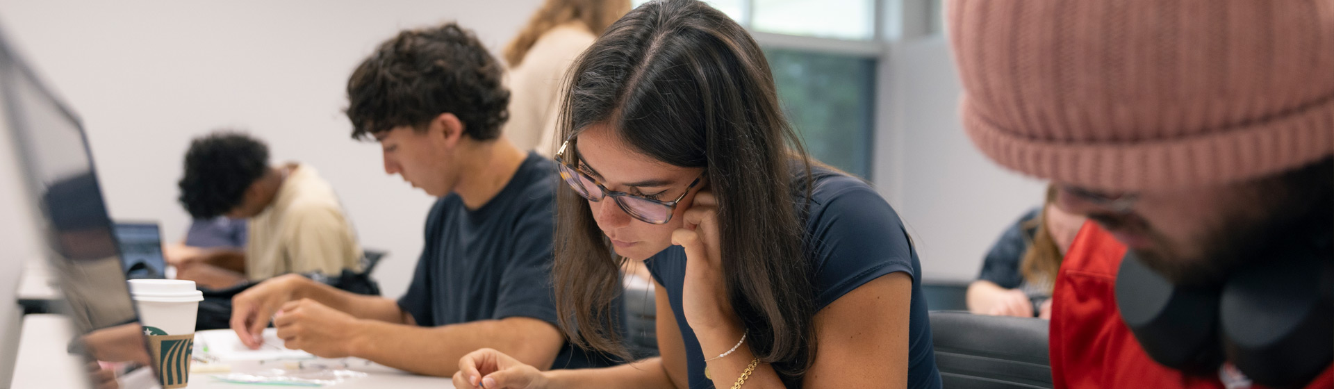 Students seated at a long table near computer monitors working on electrical boards