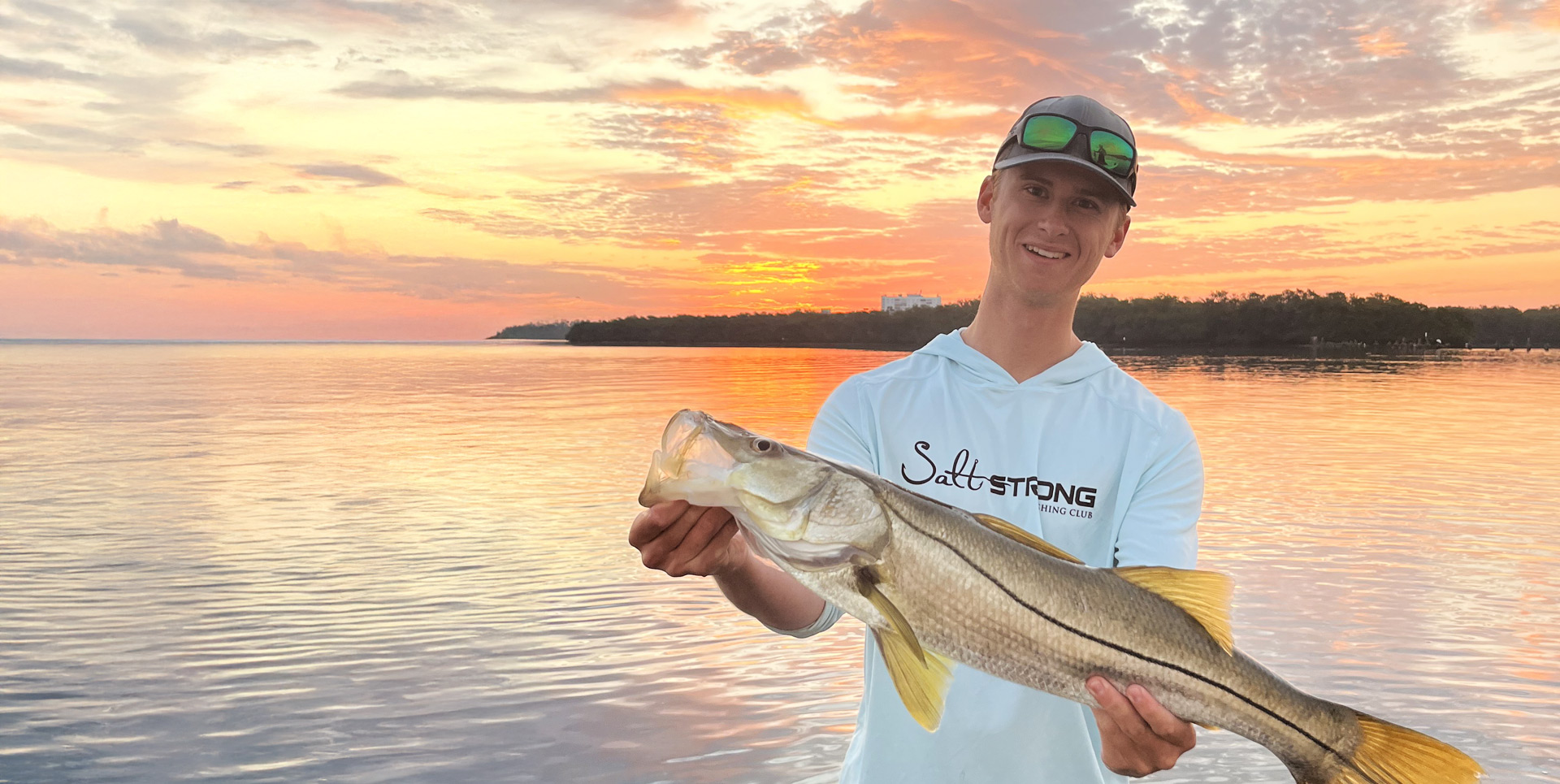Eckerd student holding a large snook as sun sets behind him across the bay