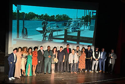 A large group of people stand on stage with image of a marina projected behind them