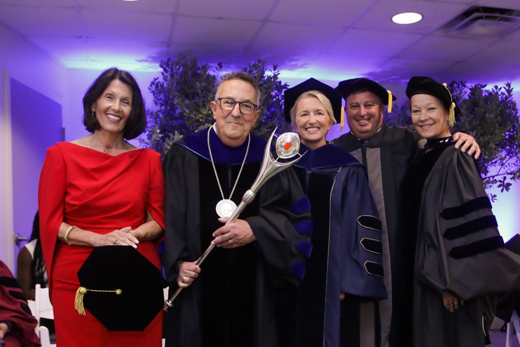 Five people stand side by side, four dressed in academic regalia, one holding a mace