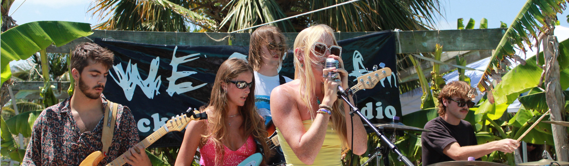 Students on outdoor stage playing instruments with a sign behind them that reads WECX