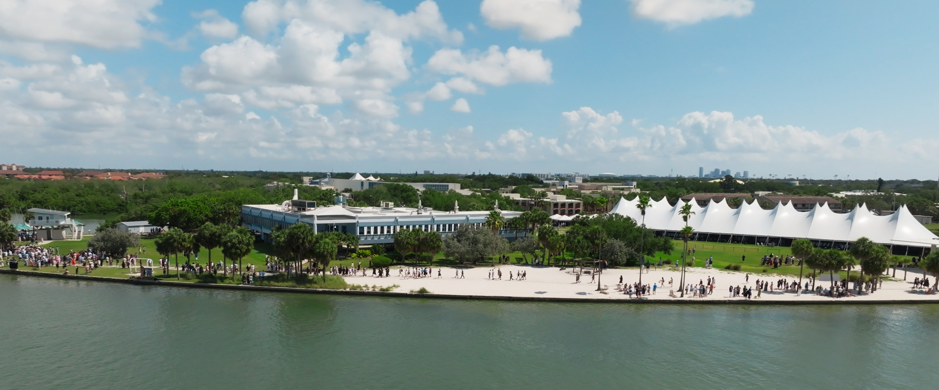 View of waterfront campus with people mingling on the beach with a very large white tent in the background